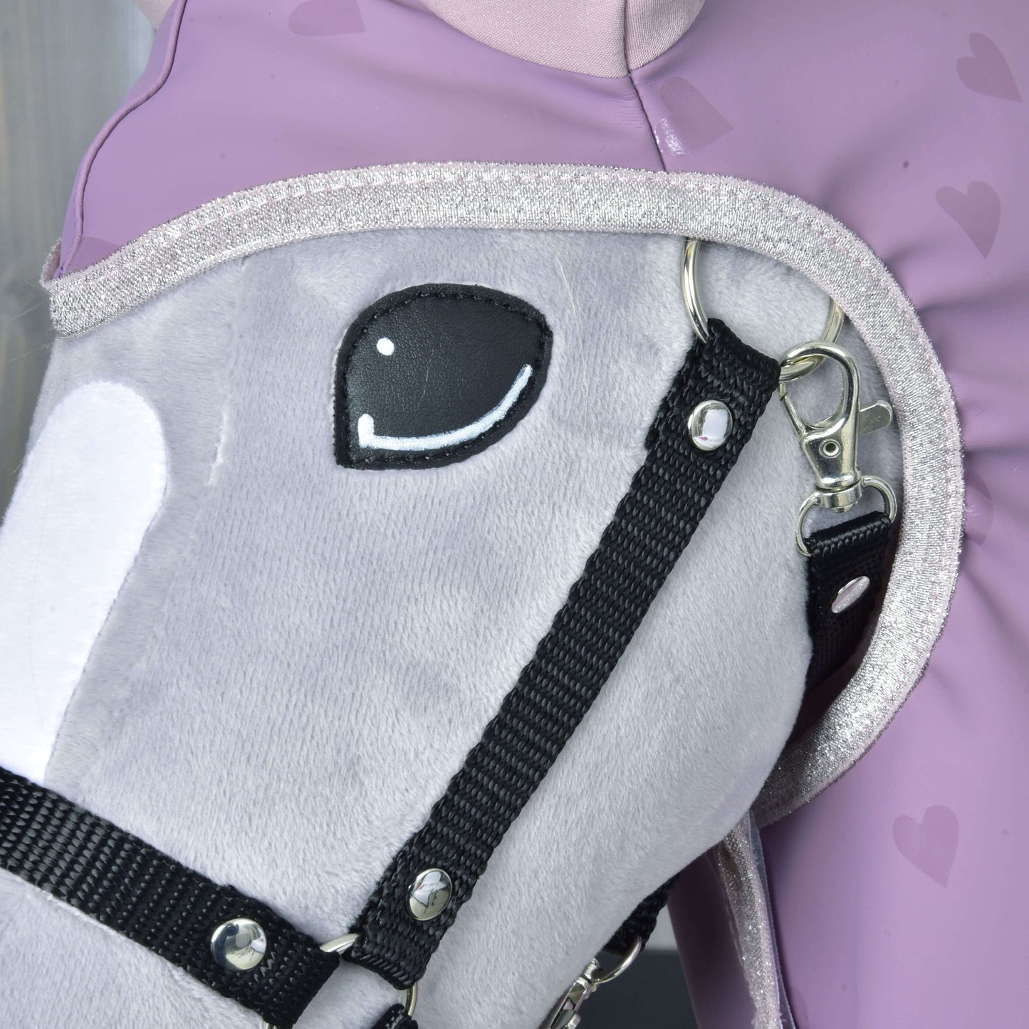 Gros plan sur un Hobby Horse gris en peluche de Hobby Horse LarDen avec bride noire, portant l’accessoire Imperméable Chéri, à côté d’une personne en tenue lavande à motif cœur. Le cheval a un œil souriant brodé et une bordure d’encolure pailletée.