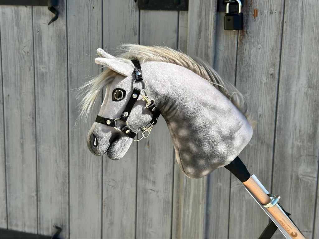 The Hobby Horse LarDen "Hobby Horse Untamed Sweety with Black Halter" is a gray plush hobby horse on a wooden stick, shown in front of a weathered wooden door with metal hinges and a padlock.