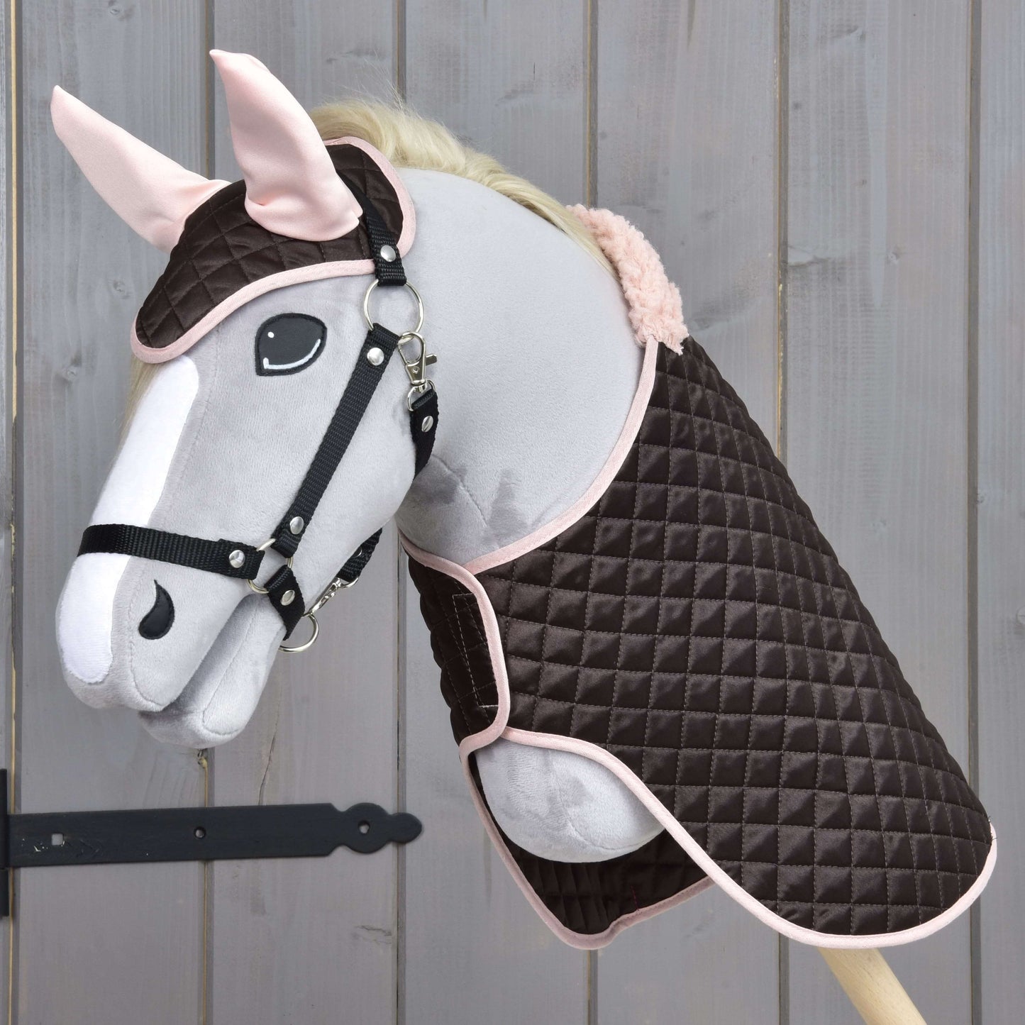 Hobby Horse LarDen Set Thermo Sweety zeigt ein plüschiges Hobby Horse mit Zaumzeug, gekleidet in eine gesteppte braun-rosa Decke mit passender Fliegenhaube, vor einem Holzhintergrund.