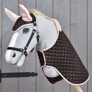 Hobby Horse LarDen Set Thermo Sweety zeigt ein plüschiges Hobby Horse mit Zaumzeug, gekleidet in eine gesteppte braun-rosa Decke mit passender Fliegenhaube, vor einem Holzhintergrund.