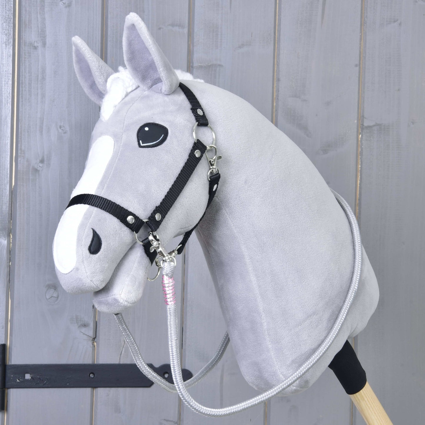 Hobby Horse LarDen Zügel Tizian zeigt ein graues Plüsch-Hobby Horse mit schwarzem Zaumzeug und handgefertigten Zügeln auf einem Holzstab, fotografiert vor hellem Holzpanelen-Hintergrund.