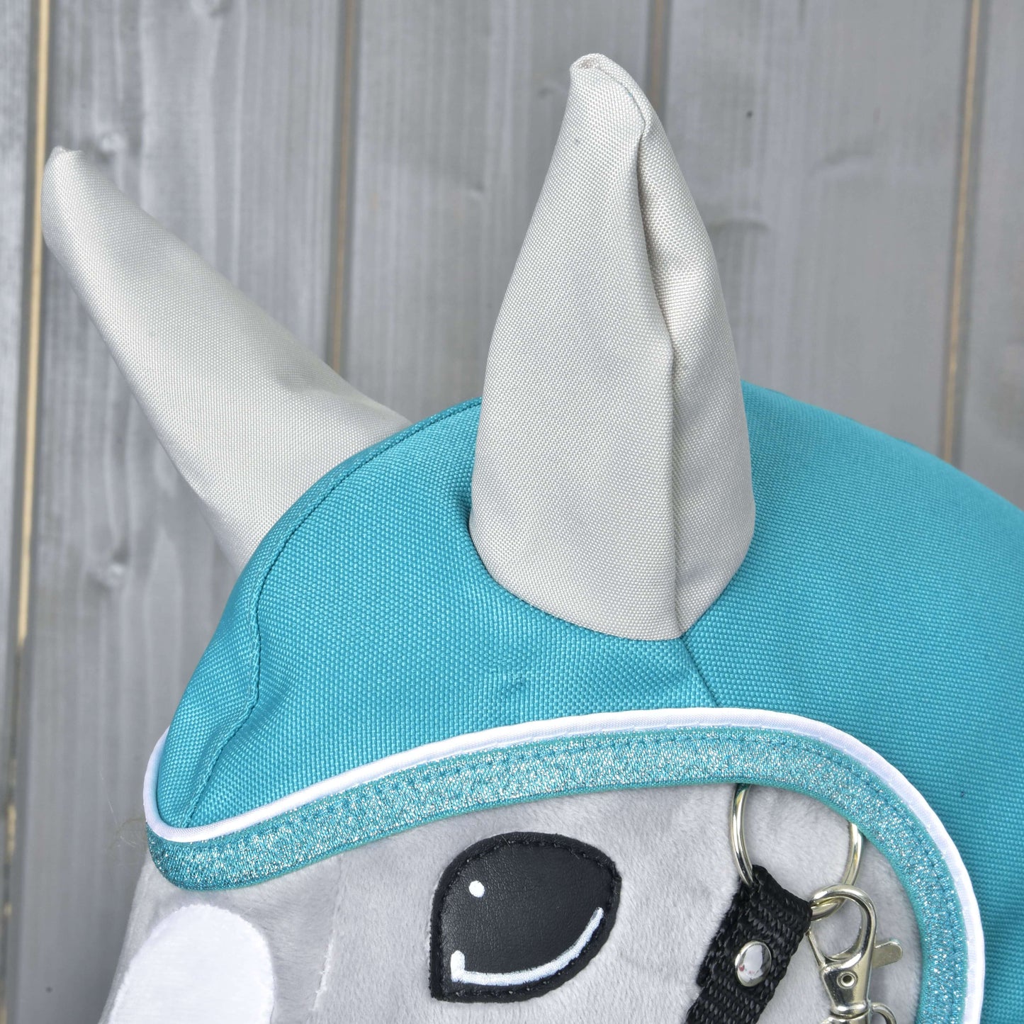 Gros plan de la tête d’un cheval en peluche Hobby Horse LarDen portant l’Imperméable turquoise, avec oreilles dressées, bordure pailletée et partie d’un licol, idéal pour jouer par tous les temps sur fond en bois.