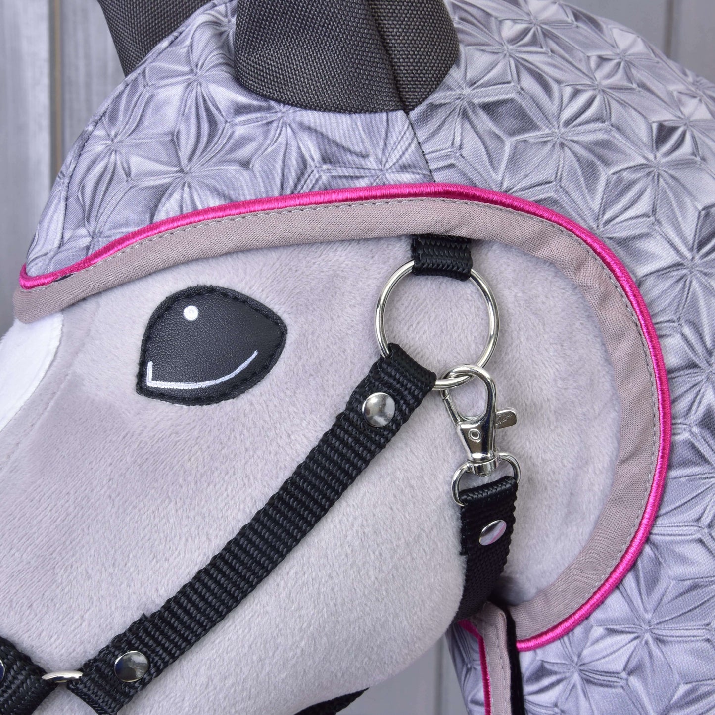 Nahaufnahme von Hobby Horse LarDen Regendecke Valentine, einem plüschigen Pferdekopf mit grauem Stoff mit geometrischem Muster, schwarzem Zaumzeug und rosa Besatz, mit einem Metallring-Clip – ein Accessoire für jedes Wetter für stilvollen Schutz.