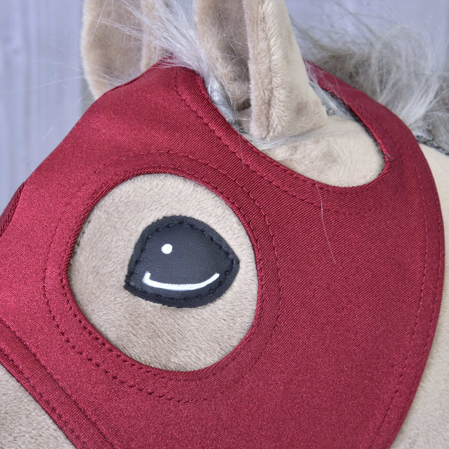 Gros plan sur le masque de cheval sans oreilles en velours de Hobby Horse LarDen, montrant une fourrure en velours beige, une bride en tissu rouge, un œil cousu noir et blanc, et une partie de la crinière grise.