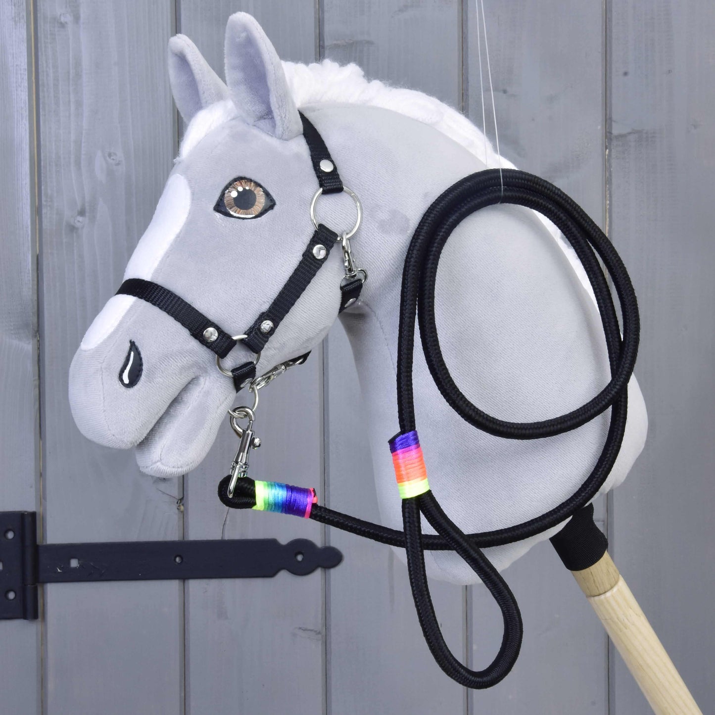 Hobby Horse - Longe arc-en-ciel de Hobby Horse LarDen, Hobby Horse gris avec crinière blanche, bride noire, rênes et longe à ruban arc-en-ciel autour du cou, présenté devant un panneau en bois.