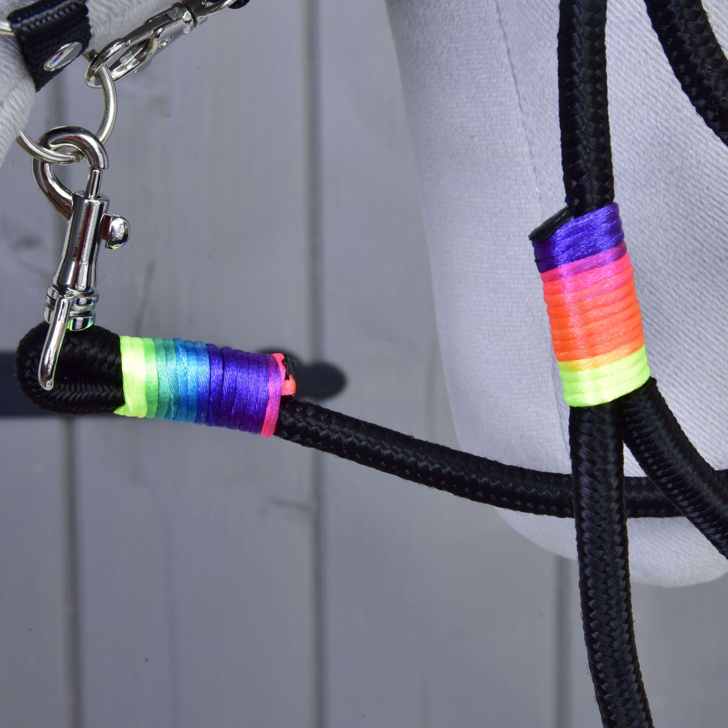 Gros plan sur la Hobby Horse - Longe arc-en-ciel de Hobby Horse LarDen, longe noire tressée avec enroulements de fil arc-en-ciel néon et fermoir argenté, attachée à un tissu gris clair, sur fond en bois.
