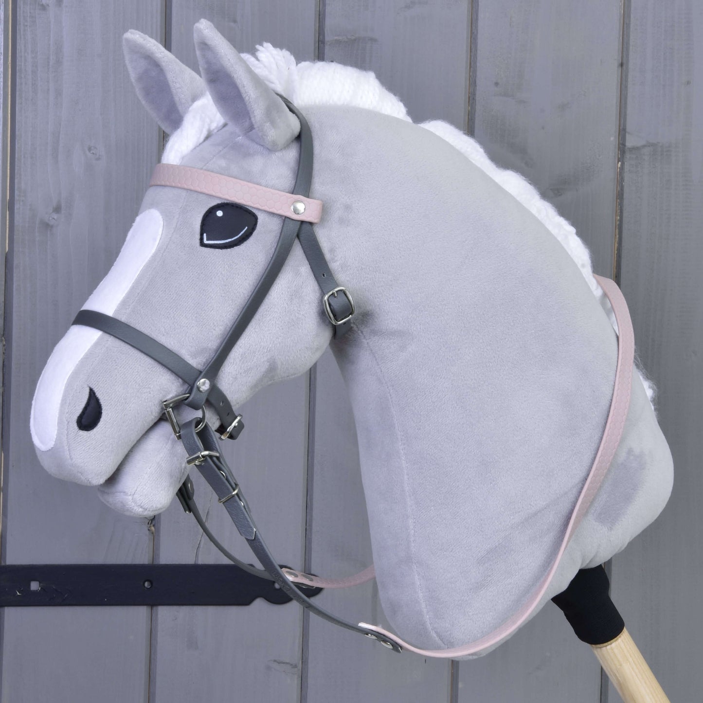 Ein graues Plüsch-Hobby Horse mit weißer Mähne und Gesichtsabzeichen trägt das Komplettes Kunstlederzaumzeug Grau mit Pink Stirnriemen von Hobby Horse LarDen, befestigt an einem Holzstab vor einer Holzpaneelwand.