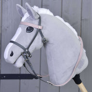 A plush gray hobby horse with a white mane and facial markings features the Hobby Horse LarDen Complete leatherette bridle grey with pink headband, attached to a wooden stick and set against a wooden panel background.