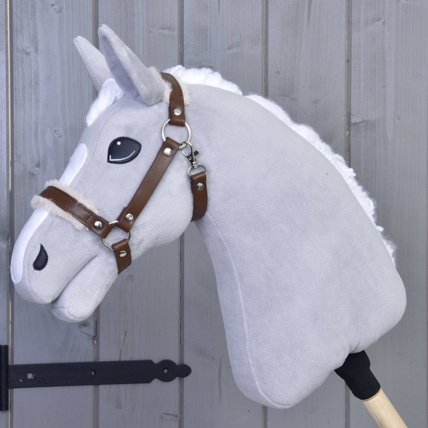 Licol avec fourrure - similicuir Noyer de Hobby Horse LarDen sur un Hobby Horse gris en peluche avec crinière blanche, licol en similicuir marron et grands yeux noirs, sur un bâton en bois appuyé contre un mur en bois gris.
