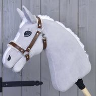 Halfter mit Fell - Kunstleder Walnut von Hobby Horse LarDen ist ein graues plüschiges Hobby Horse mit weißer Mähne, braunem Halfter aus Kunstleder und großen schwarzen Augen auf einem Holzstab, gezeigt an eine graue Holzwand gelehnt.