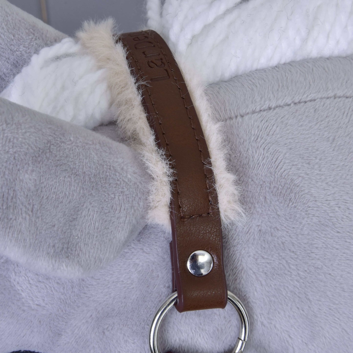 Gros plan sur le licol avec fourrure - similicuir Noyer de Hobby Horse LarDen, avec tissu en peluche gris et blanc relié par une couture duveteuse. Une sangle en similicuir marron, fixée par un bouton argenté et un anneau, traverse la jonction.