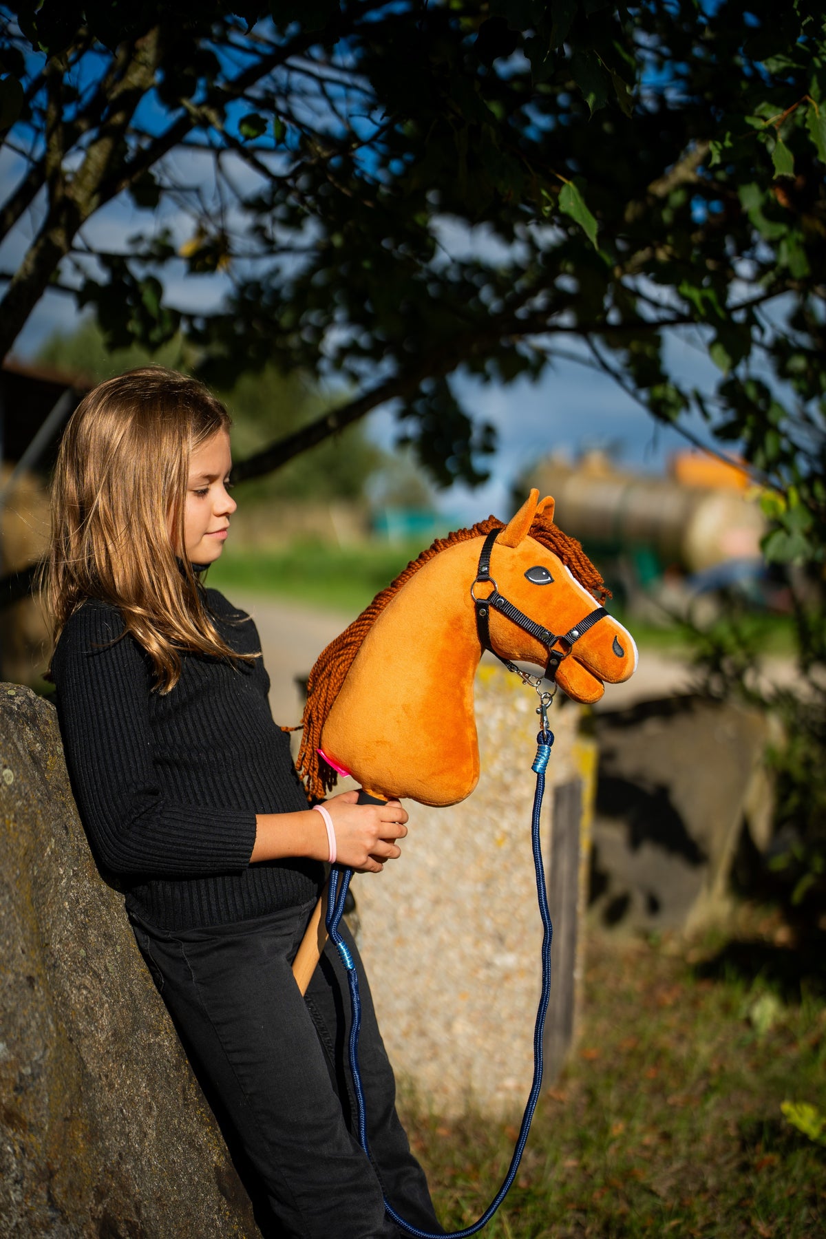 Mädchen in schwarzem Oberteil lehnt unter einem Baum an einem Steinblock und hält das Hobby Horse Wendy mit schwarzem Halfter von Hobby Horse LarDen – leuchtend heller Fuchs-Plüschkopf, rotgoldene Garnmähne – mit Blues Rope Stripe Führstrick