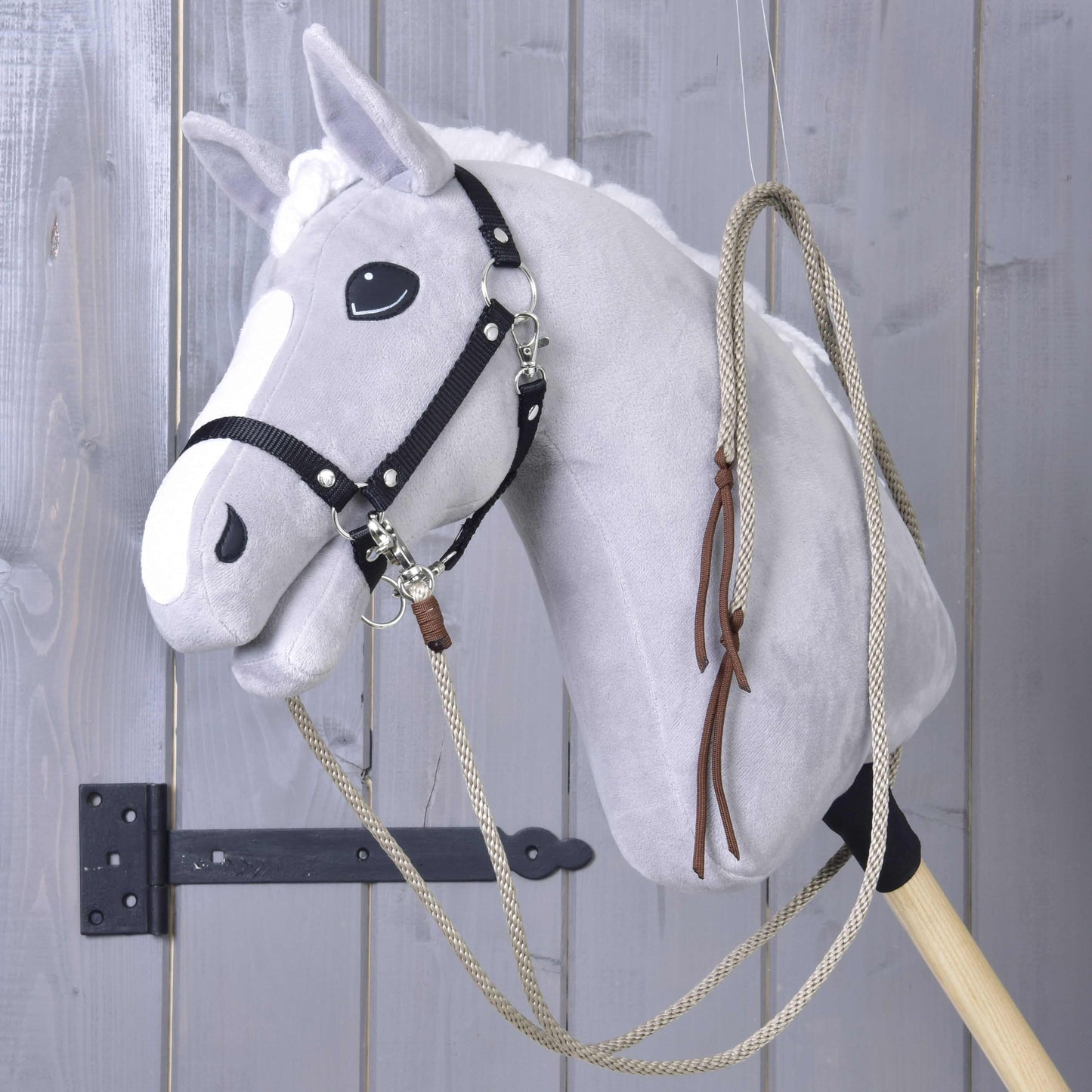 Rênes Western Fendues Dallas de Hobby Horse LarDen sur un Hobby Horse gris en peluche avec crinière blanche, harnais noir et monté sur un bâton en bois, suspendu devant un mur en panneaux de bois gris.