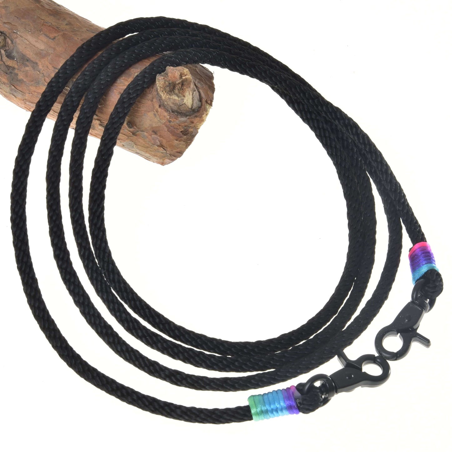 Hobby Horse LarDen Westernzügel Montana mit einer schwarzen geflochtenen Kordel mit Karabinerhaken an beiden Enden, akzentuiert mit blauen, grünen und pinken Bändern – ideal als Trainings- oder Westernzügel. Zu einem Kreis aufgewickelt vor weißem Hintergrund mit Holz oben links.