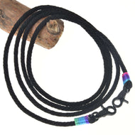 Hobby Horse LarDen Westernzügel Montana mit einer schwarzen geflochtenen Kordel mit Karabinerhaken an beiden Enden, akzentuiert mit blauen, grünen und pinken Bändern – ideal als Trainings- oder Westernzügel. Zu einem Kreis aufgewickelt vor weißem Hintergrund mit Holz oben links.