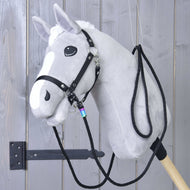 Hobby Horse LarDen Westernzügel Montana mit einem grauen Plüsch-Hobby Horse mit weißer Mähne und Blesse, schwarzem Zaumzeug mit Karabinerhaken und Westernzügeln, auf einem stabilen Holzstab montiert und vor einer grauen Holzwand präsentiert.