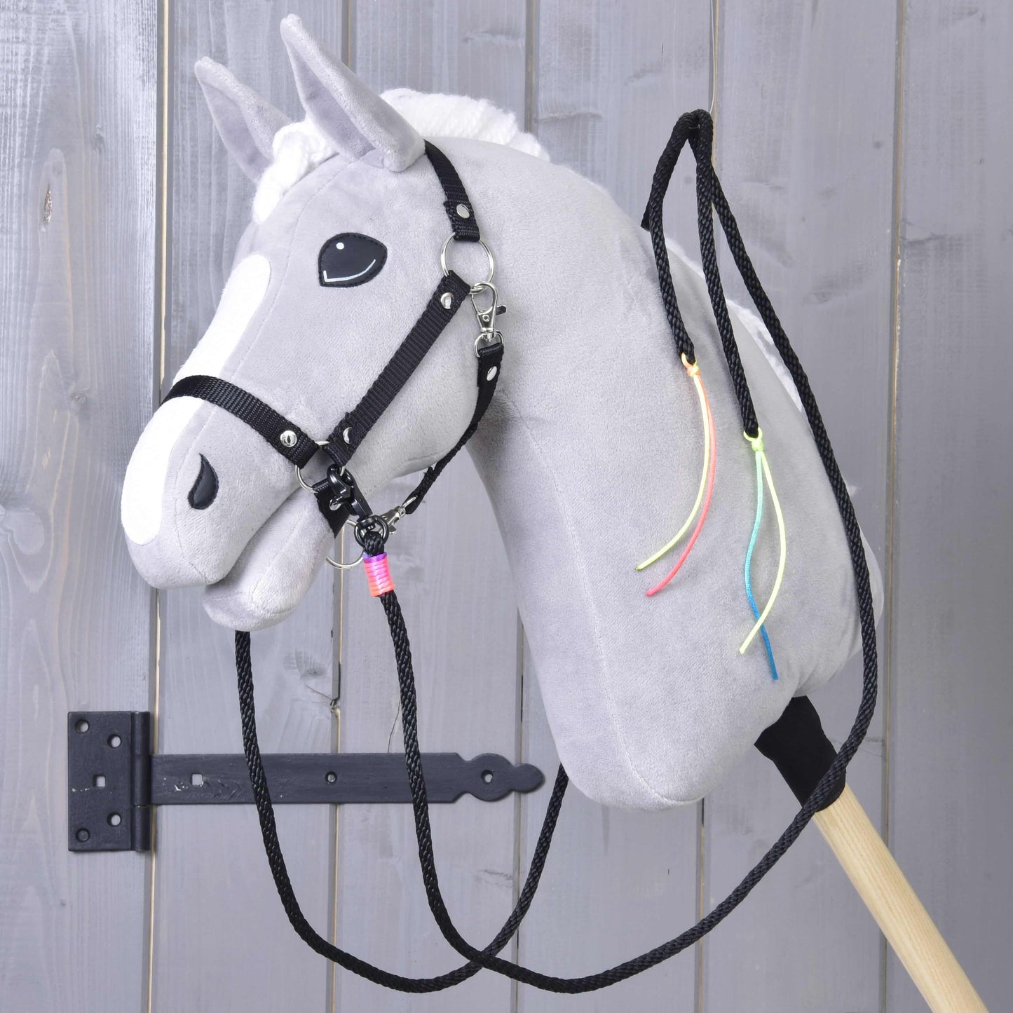 Rênes Western Fendues Texas de Hobby Horse LarDen sur un cheval en peluche gris avec crinière blanche, bride noire, rênes western fendues et rubans colorés au cou, monté sur un bâton en bois devant une porte en bois grise.