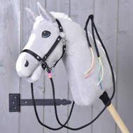 Hobby Horse LarDen Offene Westernzügel Texas mit einem grauen Plüschpferd mit weißer Mähne, schwarzem Zaumzeug, offenen Westernzügeln und bunten Halsbändern, auf einem Holzstab montiert vor einer grauen Holztür.
