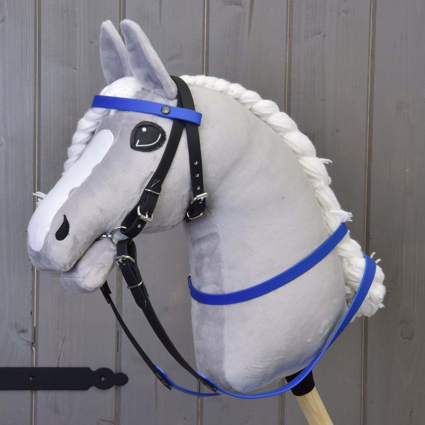 Hobby Horse en peluche de Hobby Horse LarDen avec visage gris, crinière blanche et rênes caoutchoutées bleues, monté sur un bâton en bois et placé devant un fond en panneaux de bois.