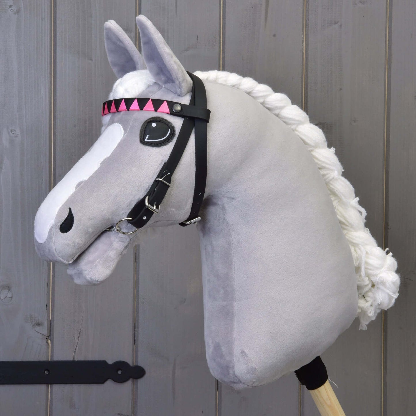 The Hobby Horse LarDen "Bridle with 2 Headbands Pink (size M)" features pink triangle accents and a bridle without noseband, shown on a grey plush hobby horse with white yarn mane and detachable reins, against a wooden background.