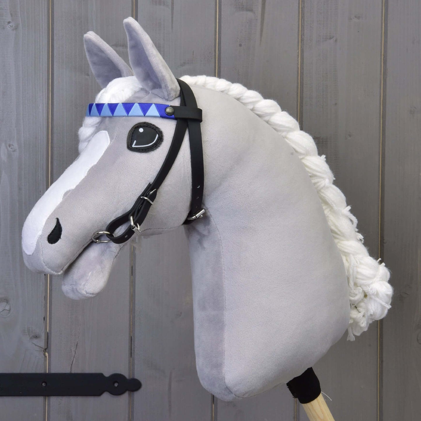 Zaumzeug mit 2 Stirnbändern Blau (Gr. M) von Hobby Horse LarDen an einem grauen plüschigen Pferd mit geflochtener weißer Mähne, schwarzem Zaumzeug, blau gemustertem Stirnband und abnehmbaren Zügeln, auf einem Holzstab vor einer Holzwand.
