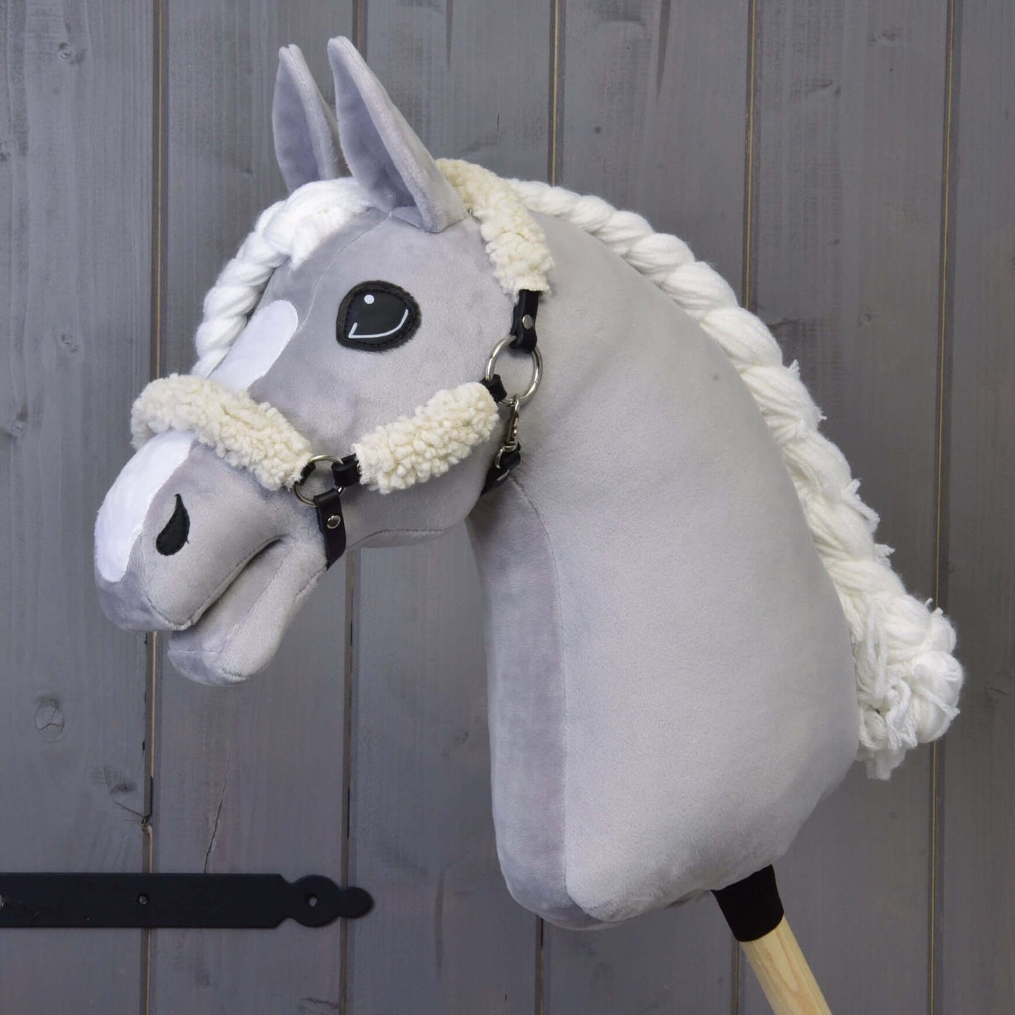 Licou noir avec fourrure complète - similicuir de Hobby Horse LarDen, avec Hobby Horse en peluche gris, licou noir entièrement doublé de fourrure avec détails en similicuir, monté sur un bâton en bois sur fond de planches en bois.