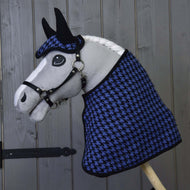 Ein Hobby Horse LarDen Hobby Horse mit weißer Mähne und schwarzem Zaumzeug trägt das „Set festlich blau kariert (Ohrenkappe + Decke)“ mit Ohrenkappe und Decke im Hahnentrittmuster und posiert vor einer grauen Holzwand.