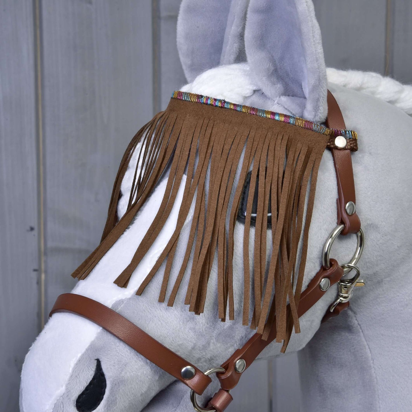Licol marron avec franges de Hobby Horse LarDen, présenté sur un Hobby Horse en peluche gris avec crinière blanche, avec franges décoratives sur le frontal, sur fond en bois.