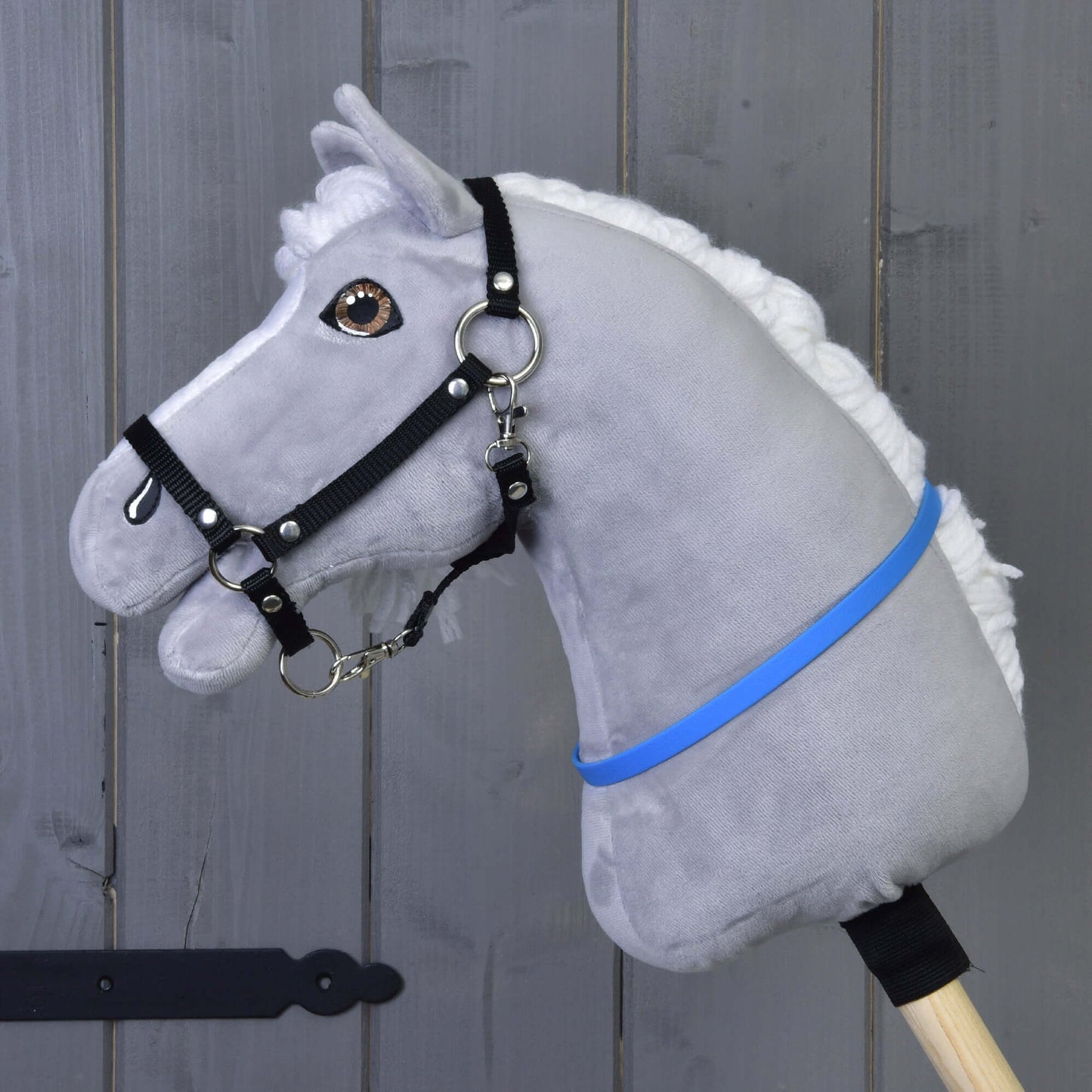 Hobby Horse cordeo bleu (taille S) de Hobby Horse LarDen, avec corps en peluche gris, crinière blanche, bride noire et ruban bleu-violet sur un bâton en bois, présenté devant un mur en bois gris.