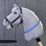 The Hobby Horse LarDen "Hobby Horse cordeo blue (size S)" features a gray plush body, white mane, black bridle, and blue-violet ribbon on a wooden stick, displayed against a gray wooden wall.