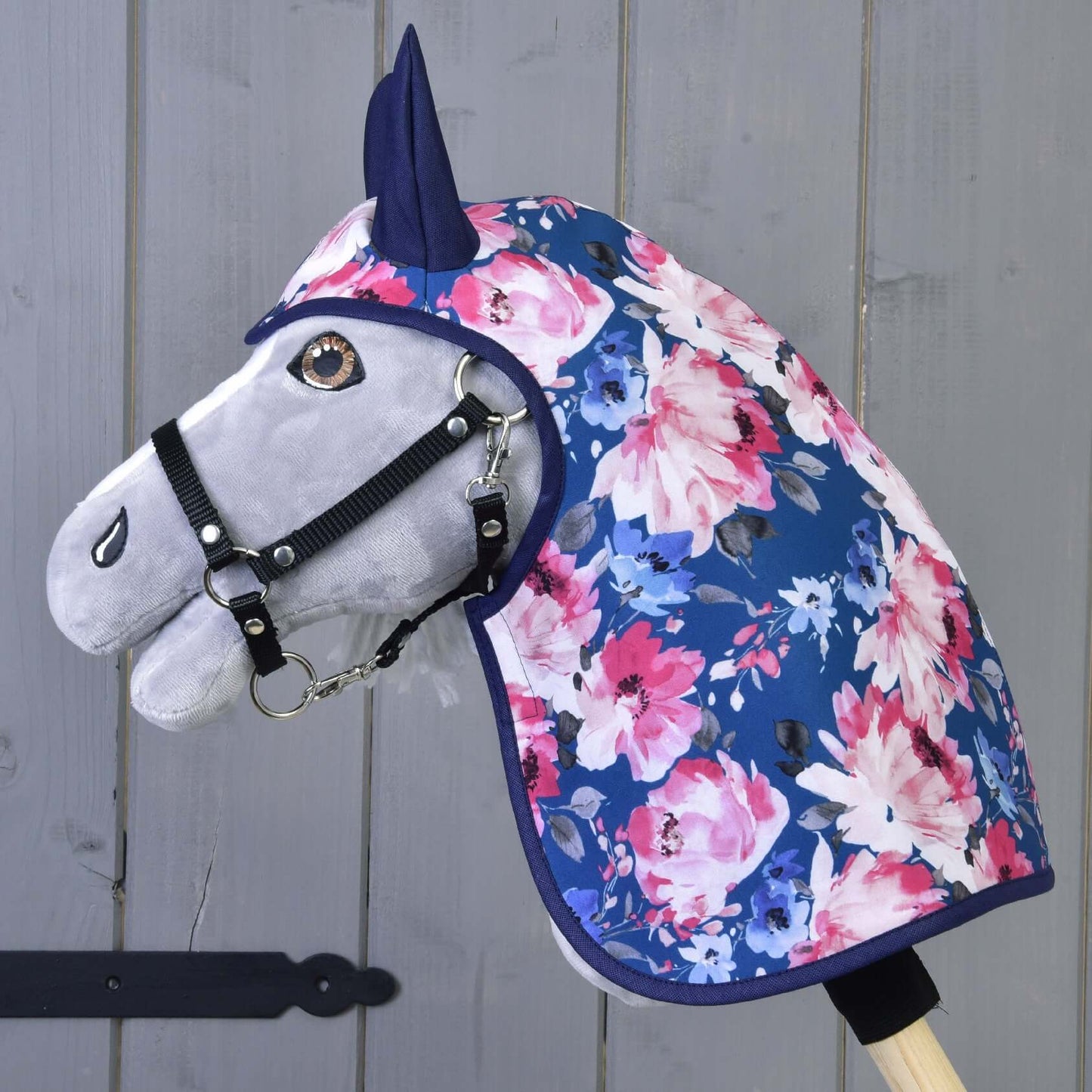 Das Hobby Horse LarDen „Regendecke Pfingstrose“ ist ein graues Hobby Horse mit schwarzem Zaumzeug, das eine blaue Regendecke mit rosa und weißen Blumen trägt, montiert auf einem Holzstab vor einer grauen Holzwand.