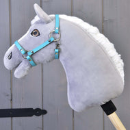 A gray plush hobby horse with a white mane and blue bridle wears the Hobby Horse LarDen Halter - black leatherette (Size S), mounted on a wooden stick in front of gray wooden panels.