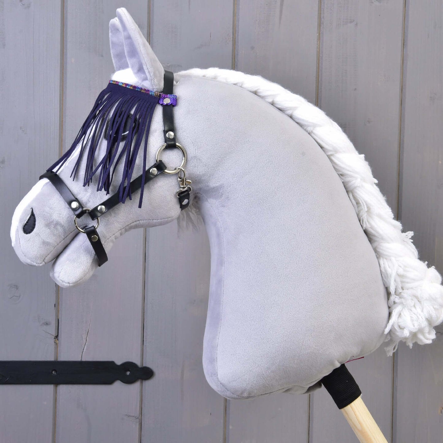 Gros plan sur un jouet Hobby Horse LarDen avec corps gris, crinière blanche en fil et licol noir avec franges lilas, sur fond en bois.