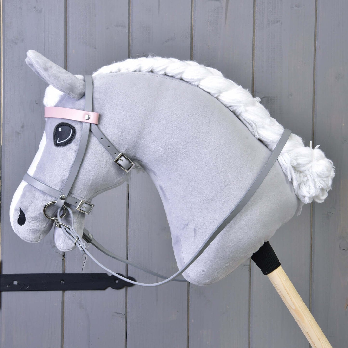 Hobby Horse en peluche gris de Hobby Horse LarDen avec crinière en fil blanc, yeux noirs et Ensemble gris en similicuir - bride, rênes et martingale, monté sur un bâton en bois sur fond de mur en bois gris.