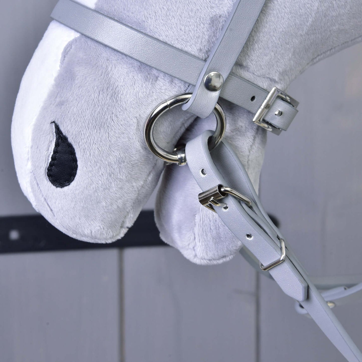 Gros plan du Hobby Horse en peluche gris de Hobby Horse LarDen portant l’Ensemble gris en similicuir - bride, rênes et martingale avec anneaux et boucles métalliques. Présente des yeux noirs cousus ; panneaux de bois flous en arrière-plan.