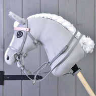 Ein graues Plüschpferd von Hobby Horse LarDen mit weißer Mähne trägt das Graues Kunstlederset - Zaumzeug, Zügel und Martingale, befestigt an einem Holzstab vor einer grauen Holzwand.